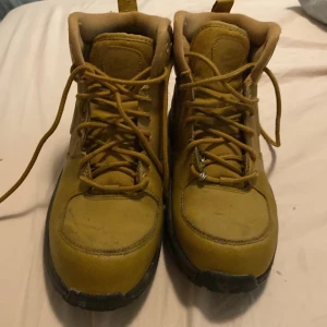 Gula boots från Nike med snörning - Säljer ett par gula boots från Nike med snörning och högt skaft. Skorna har en robust svart sula och är tillverkade i skinn. Perfekta för höst och vinter när du vill ha både stil och funktion.