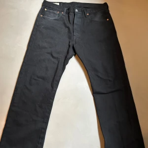 Svarta Levi's 501 jeans W32 L30 - Klassiska svarta Levi's 501 jeans med raka ben och fem fickor. Byxorna har knappgylf och den ikoniska läderpatchen bak i midjan. Perfekta för dig som gillar tidlös stil och vill ha ett par jeans som funkar till allt.