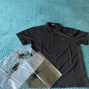 Svart pikétröja från Polo Ralph Lauren - Säljer en klassisk svart pikétröja från Polo Ralph Lauren med röd broderad logga på bröstet. Tröjan har krage, knappar och är tillverkad i bomull. Perfekt för en stilren look.