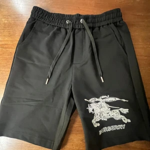 Svarta shorts från Burberry med broderad logga - Svarta shorts från Burberry med vit broderad logga och motiv på vänster ben. Shortsen har snörning i midjan och sidofickor. Perfekta för en avslappnad och stilren look. Skriv om ni har frågor