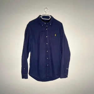 Mörkblå skjorta från Polo Ralph Lauren - Klassisk mörkblå skjorta från Polo Ralph Lauren med button-down krage och gul broderad logga på bröstet. Skjortan har långa ärmar och knäppning framtill. Perfekt för en stilren och tidlös look.