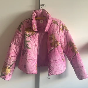 Rosa pufferjacka från Vans x Realtree - Säljer en rosa pufferjacka från Vans i samarbete med Realtree. Jackan har ett unikt camouflageinspirerat mönster med grenar och löv i grönt och gult. Dragkedja framtill och hög krage. 