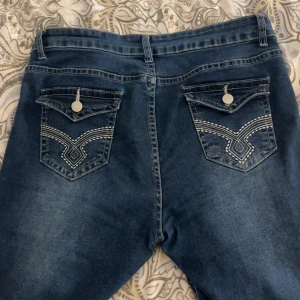 Blå jeans med broderade bakfickor - Säljer ett par blå jeans med snygga broderade detaljer och knappar på bakfickorna. Jeansen har normal passform och klassisk femficksdesign. Perfekta för dig som vill ha ett par jeans med lite extra detaljer, köpte dom för 190kr. Har används ett par gånger!
