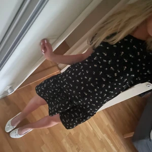 Svart blommig kortklänning - Söt svart kortklänning med små blommor i vitt och rosa. Klänningen har korta ärmar och en lätt volang nertill. Perfekt för sommarens alla tillfällen och enkel att styla med sneakers eller sandaler.