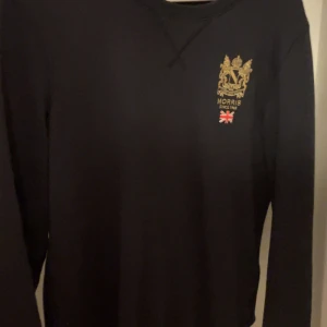 Svart långärmad tröja från Morris - Svart långärmad tröja från Morris med guldfärgat broderat emblem och liten brittisk flagga på bröstet. Klassisk och stilren design, perfekt för en snygg vardagslook.