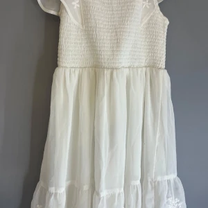 Vit somrig klänning med broderade detaljer - Söt vit klänning med smock upptill och volangärmar. Kjolen är luftig och har broderade blommor längst ner.  använd fåtal gpnger och har inga defekter, hål eller andra tecken på användning❤️