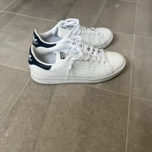 Adidas Stan Smith vita sneakers - Klassiska vita Adidas Stan Smith sneakers med mörkblå detalj på hälen och perforerade ränder på sidorna. Skorna har snörning och är tillverkade i skinn. Perfekta för en stilren och tidlös look.