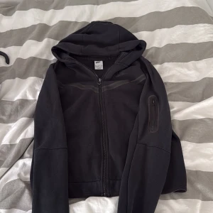 Nike tech hoodie med dragkedja (svart) - Svart hoodie från Nike med dragkedja framtill och huva. Jackan har en ficka med dragkedja på ena ärmen och är långärmad. Perfekt för en avslappnad stil.