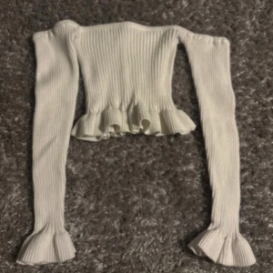 Vit ribbad croppad topp med volang - Trendig vit långärmad topp med ribbad struktur och volangdetaljer vid ärmslut och nederkant. Croppad modell som passar perfekt till höga jeans eller kjol. Snygg och stilren design.