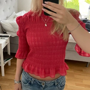 Röd volangtopp med smock - Säljer en röd topp med smock och volangdetaljer. Toppen har trekvartsärm och en lätt croppad passform. Perfekt för dig som vill ha något färgglatt och trendigt.