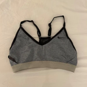 Grå sport-bh från Nike - Säljer en grå sport-bh från Nike med svarta detaljer och justerbara axelband. Bh:n har en bred, ljusgrå resår nedtill och är tillverkad i Dri-FIT-material som andas. Perfekt för träning och sportiga aktiviteter.