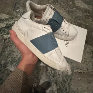 Valentino Garavani Open sneakers i vitt och blått - Säljer ett par snygga Valentino Garavani Open sneakers i vitt skinn med bred blå rem över vristen. Skorna har vita snören, rund tå och platt sula med nitar baktill. Perfekta för dig som gillar stilrena och exklusiva sneakers. Låda ingår! OBS replacement sula ingår