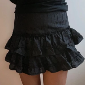 Svart volangkjol med broderade detaljer - Supersöt svart kortkjol med bred midja och flera lager volanger. Kjolen har broderade detaljer upptill och är perfekt för att skapa en trendig look. Passar dig som gillar feminina och lekfulla plagg.