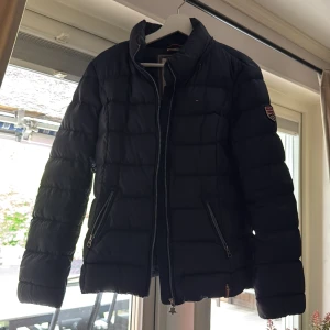Mörkblå pufferjacka från Hilfiger Denim - Säljer en mörkblå pufferjacka från Hilfiger Denim med dragkedja och två fickor framtill. Jackan har lång ärm och patchdetalj på ena ärmen. Perfekt för kyligare dagar.