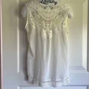 Vit ärmlös klänning med spetsdetaljer - Säljer en söt vit ärmlös klänning med vackra spetsdetaljer upptill och nertill. Klänningen har en romantisk känsla med broderad spets över axlar och rygg samt längs fållen. Perfekt för sommarens alla tillfällen!
