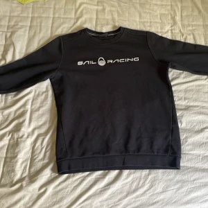 Svart sweatshirt från Sail Racing - Svart sweatshirt från Sail Racing med vit logotyp tryckt på bröstet. Klassisk rund halsringning och långa ärmar. Perfekt för en sportig och stilren look.
