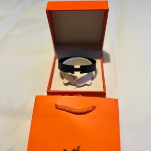 Svart armband  - Stilrent svart armband med ikoniskt H-spänne. Armbandet har en modern och minimalistisk design i svart färg och levereras i originalask. Perfekt accessoar för att lyfta din outfit. 16 cm långt.