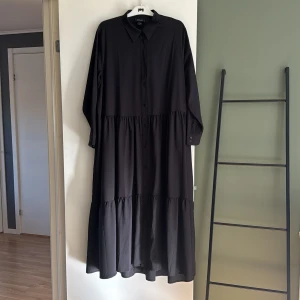 Svart långklänning med krage från Monki - Svart långklänning från Monki med klassisk skjortkrage och knappar framtill. Klänningen har lång ärm och är sydd i flera volanger för en luftig känsla. Perfekt för dig som gillar en enkel men stilren look.