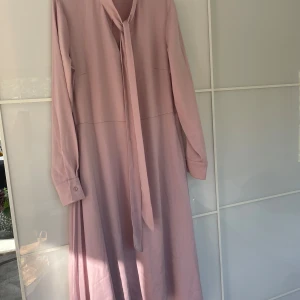 Ljusrosa långklänning med knytband - Säljer en ljusrosa långklänning med lång ärm och snyggt knytband vid halsen. Klänningen har en rak och lös passform som ger en avslappnad men stilren look. Perfekt för dig som gillar enkel och elegant design.