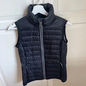 Svart dunväst från Berkeley - Snygg svart dunväst från Berkeley med hög krage och dragkedja framtill. Västen har två fickor med dragkedja och quiltad design. Perfekt att slänga över en hoodie eller tröja för extra värme.