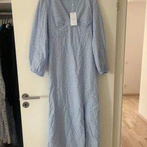 Ljusblå långklänning från Vila - Superfin ljusblå långklänning från Vila med v-ringning och broderade blommor. Klänningen har långa puffärmar och en elegant, feminin siluett. Perfekt för sommarens speciella tillfällen!