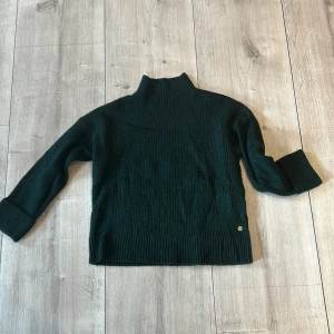 Mörkgrön stickad polotröja - Mysig mörkgrön stickad polotröja med ribbade muddar och hög krage. Perfekt för kyliga dagar och enkel att matcha med olika outfits. Klassisk och stilren design utan synliga detaljer framtill.