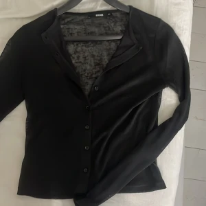 Svart transparent kofta från BikBok - Säljer en svart, tunn och transparent kofta från BikBok i storlek XS. Koftan har långa ärmar och knäppning framtill med svarta knappar. Perfekt att styla över ett linne eller topp för en trendig look.