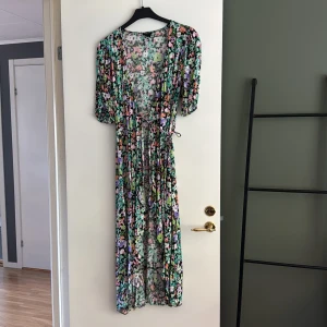 Blommig långklänning med knytband - Säljer en långklänning med färgglatt blommigt mönster i grönt, lila, rosa, orange och svart. Klänningen har korta ärmar, v-ringning och knytband i midjan för en snygg passform. Perfekt för sommarens alla tillfällen!