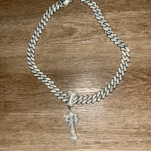 Iced ut trapstar kedjehalsband med korshänge - Säljer ett chunky halsband i silverfärg med glittriga stenar och ett stort korshänge. Kedjan är bred och täckt av stenar, och låset är också dekorerat. Perfekt statement piece för dig som gillar att sticka ut.