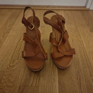Bruna kilklackade sandaler med korksula - Säljer ett par bruna sandaler med kilklack och korksula. Skorna har remmar i brunt syntetmaterial och en öppen tå. Perfekta för varmare dagar och ger en snygg höjd med stabilitet.