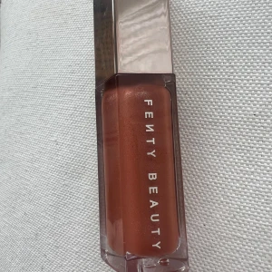 Fenty Beauty Gloss Bomb Universal Lip Luminizer 01 Fenty Glow - Superglansigt läppglans från Fenty Beauty i nyansen 01 Fenty Glow. Kommer i en snygg, femkantig förpackning med silvrigt lock. Ger en varm, rosabrun ton med shimmer och en fyllig look till läpparna.Kommer utan förpackning, oanvänd och otestad. Rensning inför flytt
