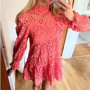 Rosa klänning  - Superfin rosa kortklänning med broderade hålmönster och volangdetaljer. Klänningen har lång ärm med resår vid ärmslut och hög hals. Perfekt för dig som vill sticka ut med en färgglad och lekfull look. Storlek s/m som nyskick