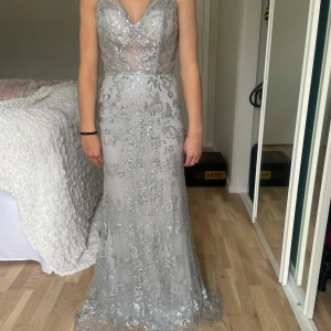 Silverglittrig långklänning från Pink Boom - Säljer en helt ny fantastisk silverfärgad långklänning från Pink Boom med v-ringning och tunna axelband. Klänningen är täckt av glittriga paljetter och har ett elegant mönster samt markerad midja. Perfekt för bal eller festkvällar! Innan min dotter valde en annan balklänning sydde en skräddare upp den o gjorde den något mindre runt midjan. Så det är nu mer en xs än s. Men oanvänd. Mått byst 35 cm rakt över, midja 31 cm. Min dotter är ca 160 cm lång. Hon har inga skor på sig på bilden.