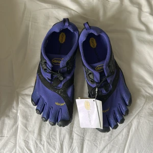 Vibram FiveFingers lila blå tåskor - Säljer ett par lila blå Vibram FiveFingers med svarta detaljer och gul logga. Skorna har individuell tådesign, snörning och grov sula för extra grepp. Perfekta för dig som gillar barfotakänsla vid träning eller utomhusaktiviteter. Eller bara vill pimpa till din vardagslook. 