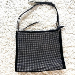 Svart axelväska med glittriga detaljer - Säljer en svart axelväska med glittriga silverdetaljer på framsidan. Väskan har justerbart axelband och stängs med dragkedja. Perfekt för att addera lite extra bling till din outfit!