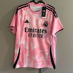 Rosa Real Madrid fotbollströja Adidas - Snygg rosa Real Madrid fotbollströja från Adidas med svart logga och klubbmärke på bröstet. Tröjan har korta ärmar, svarta ränder på axlarna och subtilt mönster i vitt och rosa. Tillverkad i lätt och ventilerande Aeroready-material.