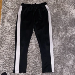 Svarta mjukisbyxor Nominal med vita ränder - Svarta trackpants. Storlek S Snygga och bekväma byxor med vit sidopanel. Resår i midjan med dragsko. 