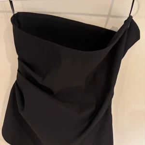 Svart asymmetrisk topp från Zara - Säljer en svart topp från Zara med cool asymmetrisk design och draperade detaljer framtill. Toppen är axelbandslös och har en stilren, modern look som sticker ut. Perfekt för dig som gillar trendiga och unika plagg.