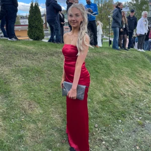 Röd axelbandslös långklänning satin - Säljer en snygg röd axelbandslös långklänning i glansigt satinmaterial. Klänningen har en figurnära passform och slät siluett, perfekt för bal eller student. Enkel och elegant design som ger en lyxig känsla.