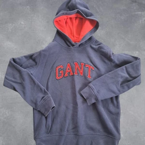 GANT hoodie - Snygg mörkblå hoodie från GANT med röd insida på huvan och broderad GANT-logga i rött framtill. Klassisk passform med långa ärmar och mjukt material, perfekt för chill dagar eller när du vill ha en sportig look.