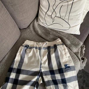 Burberry shorts - Snygga shorts från Burberry i klassiskt rutigt mönster i vitt, blått och grått. De har elastisk midja med snörning och Burberry-logga på ena benet. Perfekta för sommaren och ger en clean, lyxig vibe till din outfit.