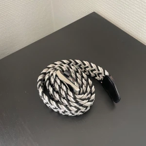 Svart och vit flätad bälte - Snyggt flätat bälte i svart och vitt med silvrigt spänne. Bältet har en cool randig look och är gjort i ett mjukt, flexibelt material med svarta skinn-detaljer vid ändarna. Perfekt för att ge outfiten lite edge.