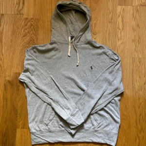 Grå hoodie från Polo Ralph Lauren - Snygg grå hoodie från Polo Ralph Lauren i storlek L med klassisk broderad logga på bröstet. Tröjan har huva med vita snören, känguruficka och är tillverkad i mjuk bomull. Tyget är rätt tunt till skillnad från vanliga hoodies så denna är perfekt nu inför sommaren!🤩 