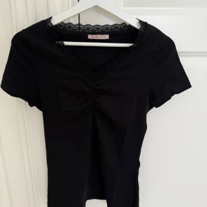 Svart topp/tshirt med spets  - Svart topp från Anna Field med korta ärmar och v-ringning dekorerad med fin spetskant. Toppen har rynkdetalj framtill som ger en snygg form. Perfekt att styla med jeans eller kjol för en trendig look. Knappt använd!