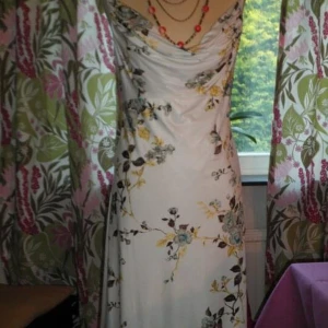 Vit långklänning med blommigt mönster - Supersnygg långklänning i vit bas med blommigt mönster i gult, blått och grått. Klänningen har draperad urringning och smal passform som framhäver figuren. Perfekt för sommarens alla tillfällen!