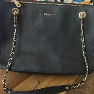 Svart axelväska från DKNY med kedja - Snygg svart axelväska från DKNY i skinn med guldfärgade kedjedetaljer och dragkedja upptill. Väskan har ett rymligt foder med DKNY-logga och flera innerfack, perfekt för att hålla ordning på dina grejer. Klassisk och stilren design med exklusiv känsla.Mått: 34*12*24cm