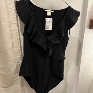 Svart volangbody från H&M - Supersnygg svart body från H&M med volangdetaljer längs axelbanden och över bysten. Prislappen kvar, aldrig använd! Perfekt för fest💛