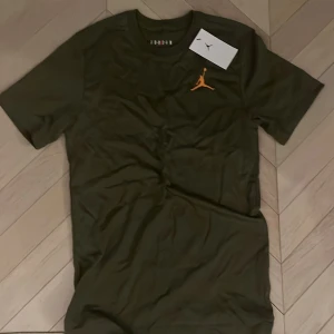 Olivgrön Jordan t-shirt med logga - Kan sänka pris vid snabb affär!     Snygg olivgrön t-shirt från Jordan med klassisk orange Jumpman-logga broderad på bröstet. T-shirten har rund hals och korta ärmar, perfekt för en sportig och avslappnad stil. Tillverkad i mjuk bomull för maximal komfort. 