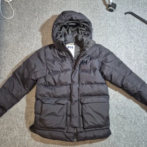 Svart pufferjacka från Helly Hansen - Svart pufferjacka från Helly Hansen med stor huva och två rejäla fickor framtill. Jackan har Helly Hansen-logga på bröstet och dragkedja hela vägen. Perfekt för kalla dagar när du vill hålla värmen och ändå se clean ut.