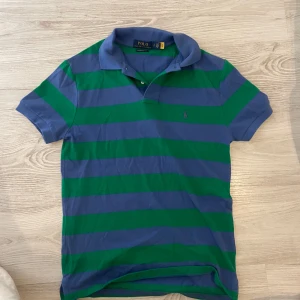 Randig pikétröja från Polo Ralph Lauren - Snygg blå och grön randig pikétröja från Polo Ralph Lauren i modellen Custom Slim Fit. Klassisk krage, korta ärmar och broderad logga på bröstet. Tillverkad i mjuk bomull, perfekt för en chill och stilren look.